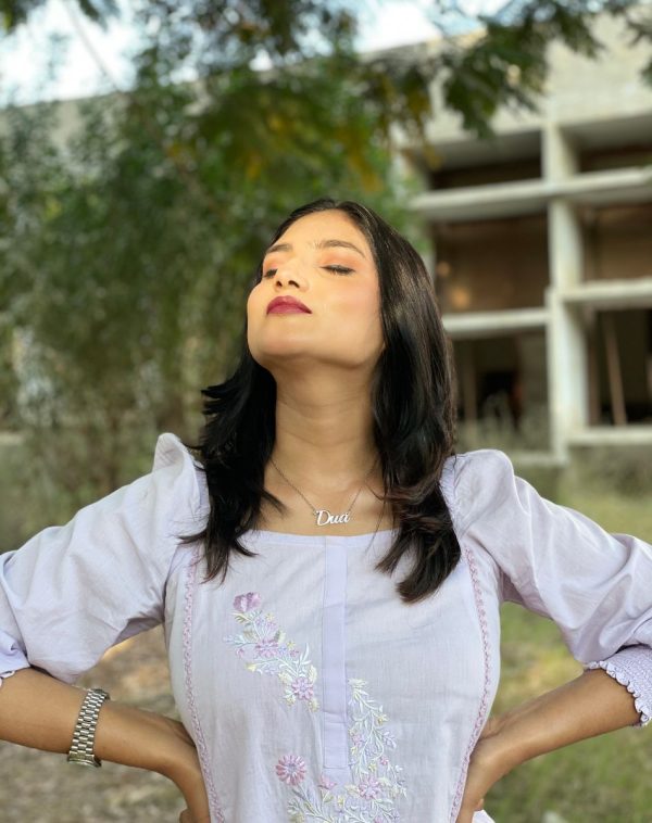 Dua wearing Single Name Silver-Plated Necklace, dressed in a lilac square-neck dress, standing outdoors with closed eyes and hands on waist, against a lush green backdrop.