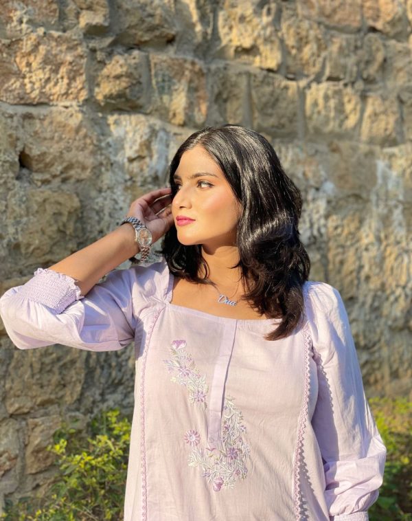 Dua wearing Single Name Silver-Plated Necklace, looking sideways with open eyes, posing with her hand behind her ear, wearing a lilac dress with square neck and puffy shoulders, against a brick wall and grassy background in sunlight.