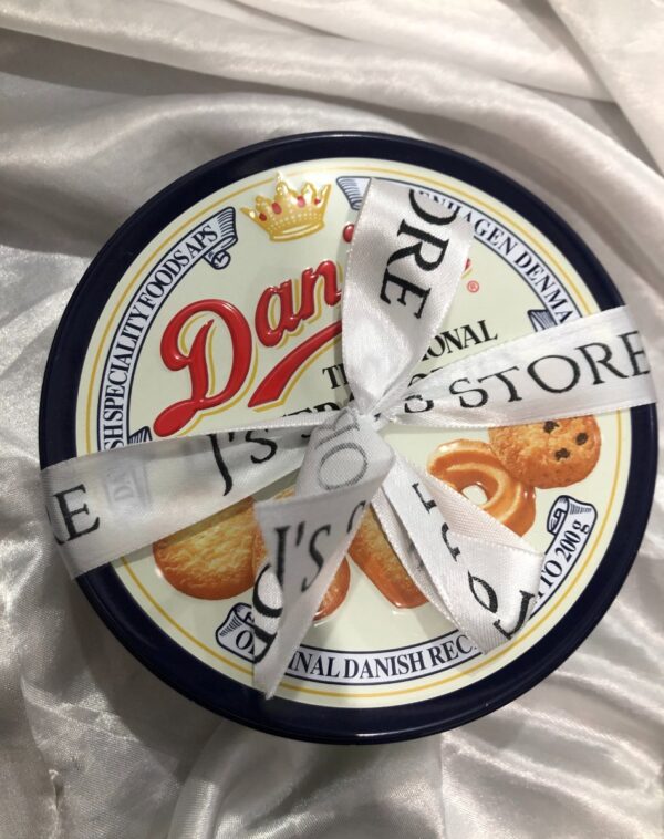 Danisa Cookies: A round, blue and white tin of Danisa Traditional Butter Cookies. The lid features the red "Danisa" logo with a crown, images of butter cookies, and text indicating "Traditional Butter Cookies" and "Original Danish Recipe." A white ribbon with black text that includes "J's STORE" is tied around the tin.