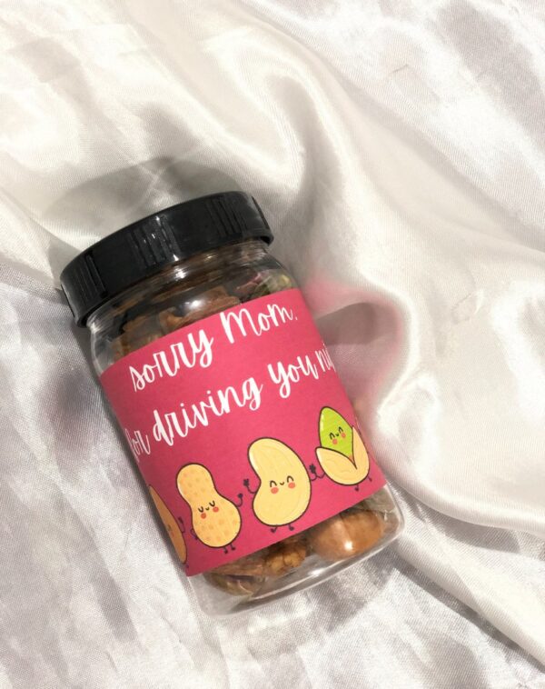 Dry Fruits Jar For Mother's Day: A clear plastic jar with a black lid, filled with a mix of dry fruits. A pink label wraps around the jar with the handwritten text "Sorry Mom for driving you nuts!" in white. Below the text are cartoon illustrations of peanuts and a pistachio with cute faces.