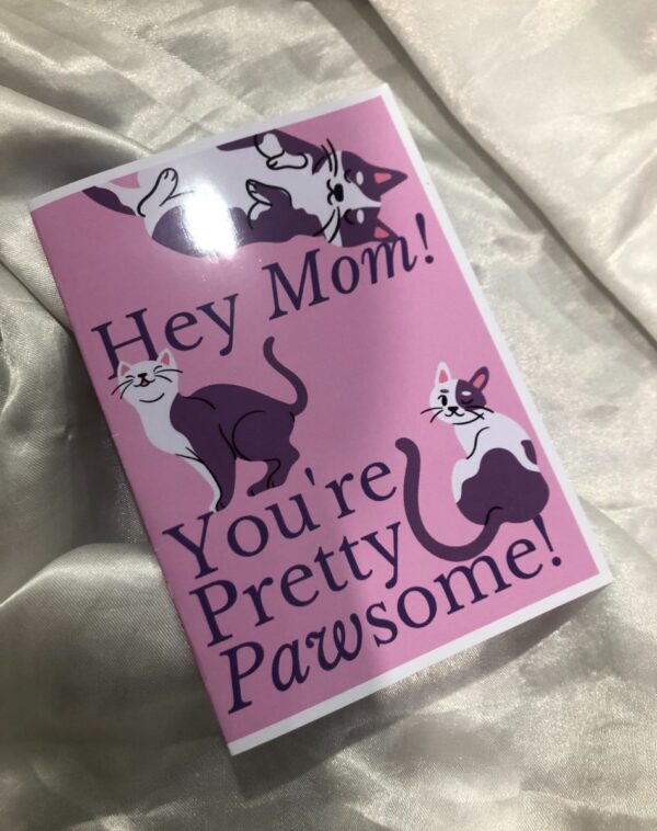 Mother's Day Greeting Card: A pink rectangular card with the text "Hey Mom!" at the top in a dark purple, playful font. Below, in a larger, also dark purple font, it says "You're Pretty Pawsome!". The card features cartoon illustrations of three cats: one black and white cat lounging upside down at the top, a solid dark purple and white cat looking to the right in the middle left, and a white cat with dark purple patches looking upwards on the middle right. This is a Mother's Day Greeting Card.