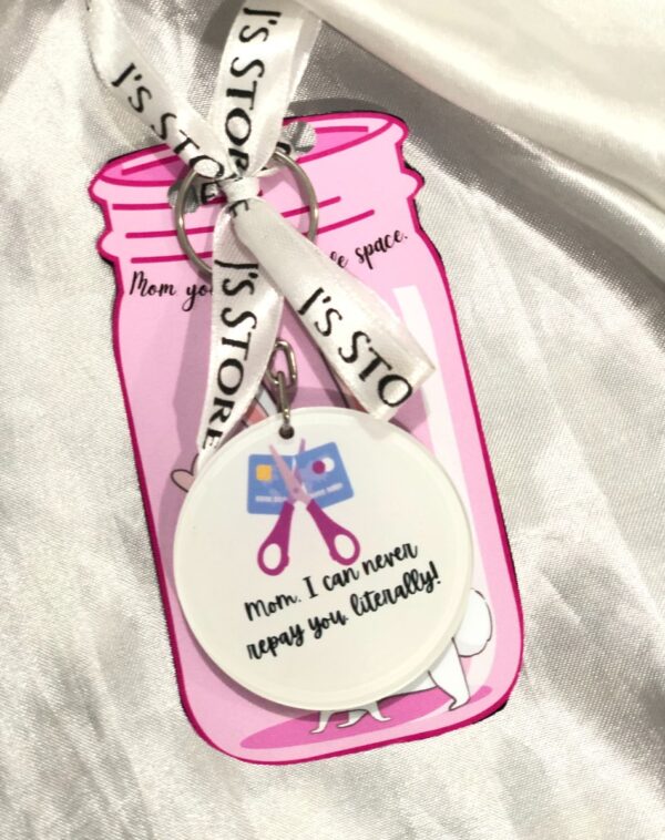 Mother's Day Keychain: A round, white keychain with scissors cutting a credit card, and the text "Mom. I can never repay you literally!". It hangs from a pink tag shaped like a jar, with the text "Mom you" and "J's STORE" on it. A white ribbon with "J's STORE" is attached to the keychain.