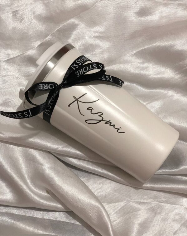 A white customized coffee tumbler with the name 'Kazmi' in black cursive. A black ribbon from J's Store is tied around the tumbler.