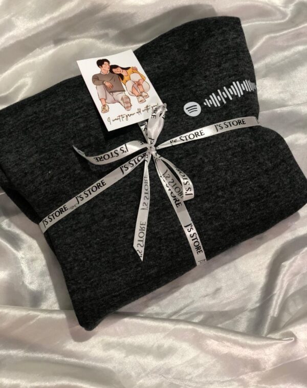 A grey hoodie with a scannable Spotify code on the sleeve. A card with a romantic message is attached. The hoodie is tied with a J's Store ribbon.