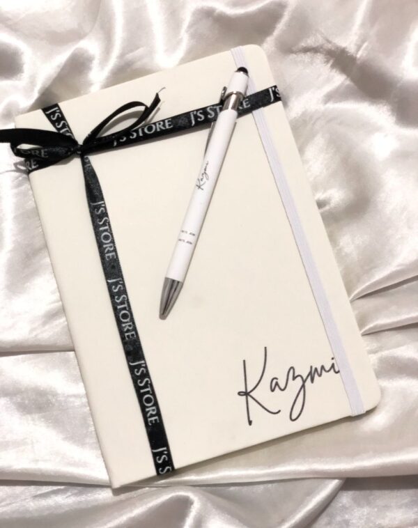 A white leather notebook and a matching pen. The notebook is personalized with the name 'Kazmi' and tied with a black ribbon.