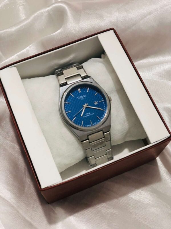 A top-down view of a Tissot PRX Powermatic 80 luxury watch featuring a textured blue "waffle" dial, silver hands, and a date window. The watch is mounted on a white display cushion inside a branded box, showcasing its integrated stainless steel bracelet and brushed metal finish.